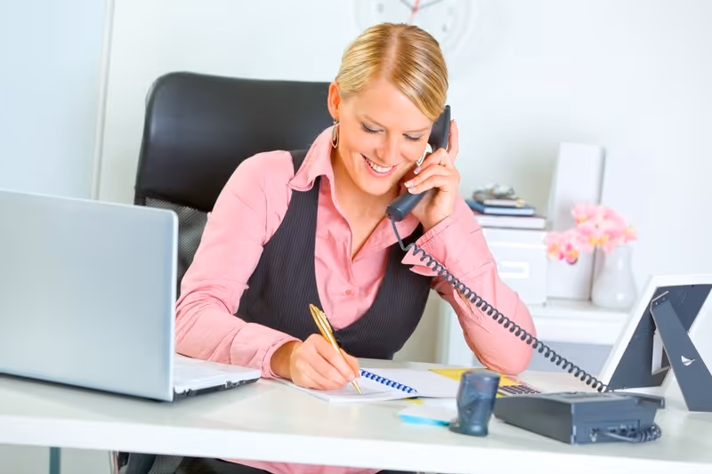 Woman in a professional setting using a telephone and taking notes, representing virtual assistant call answering services for effective customer communication.