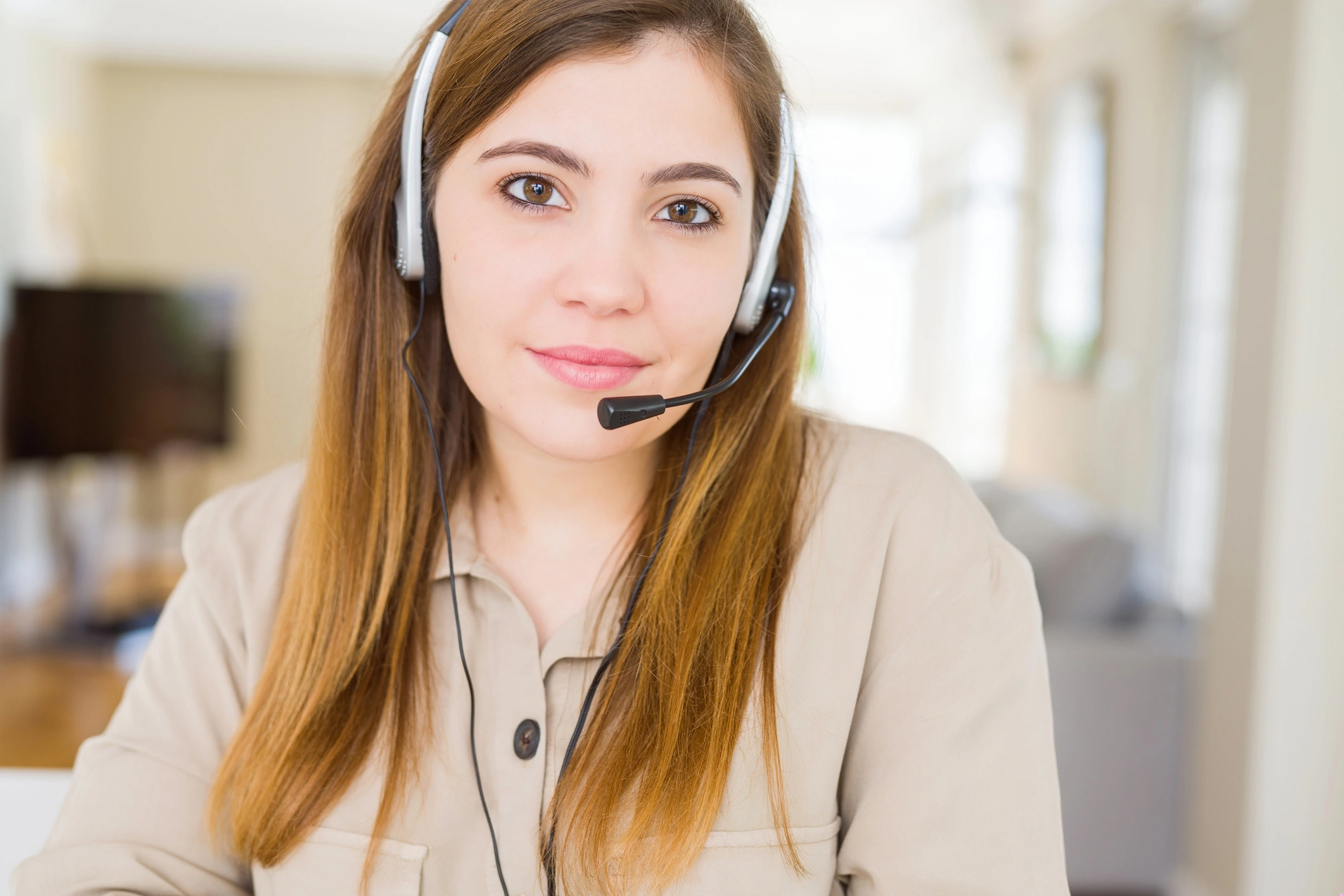 Young woman with long hair wearing a headset and beige top, sitting in a bright room.