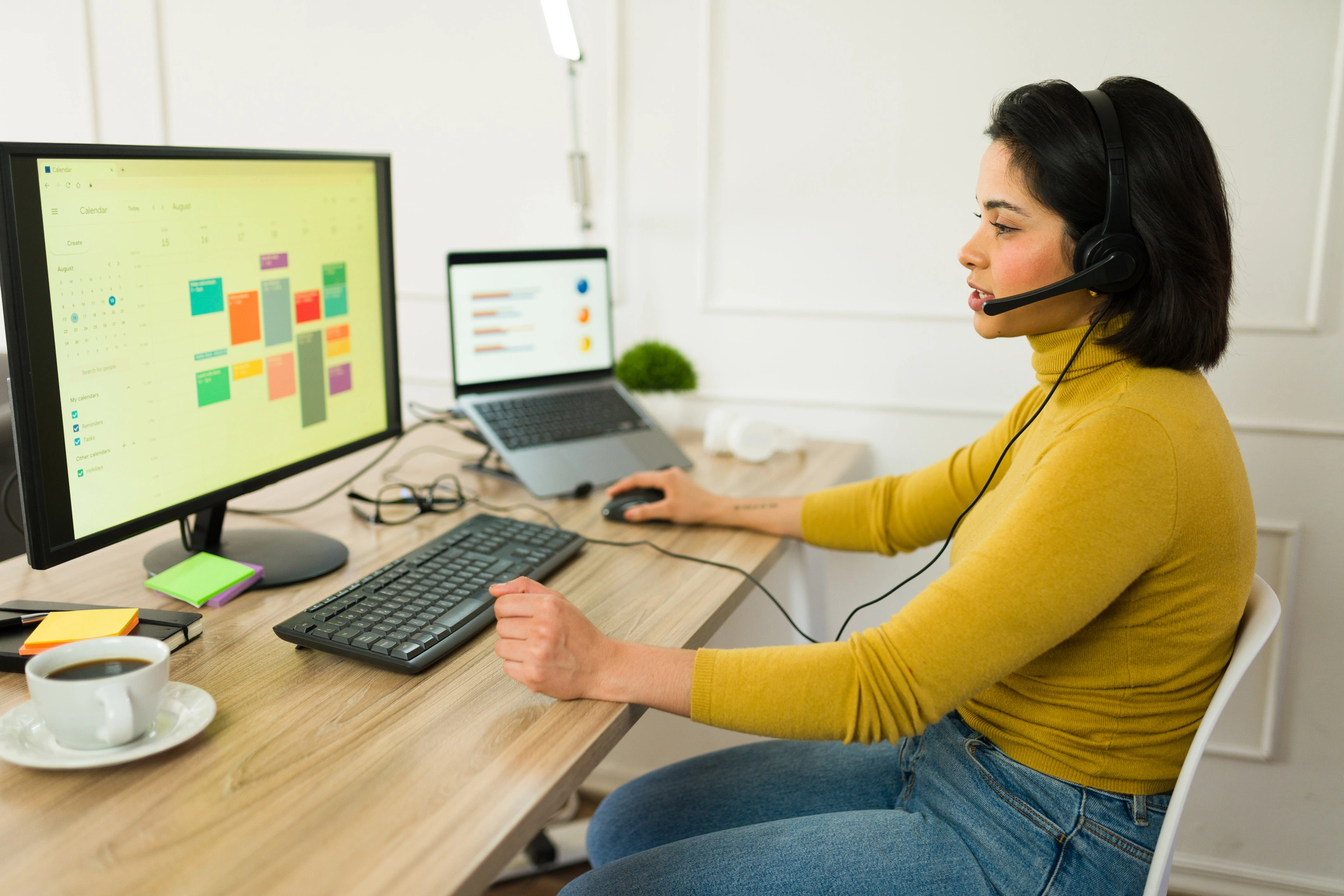 A woman in a mustard sweater works at a desk with dual monitors.