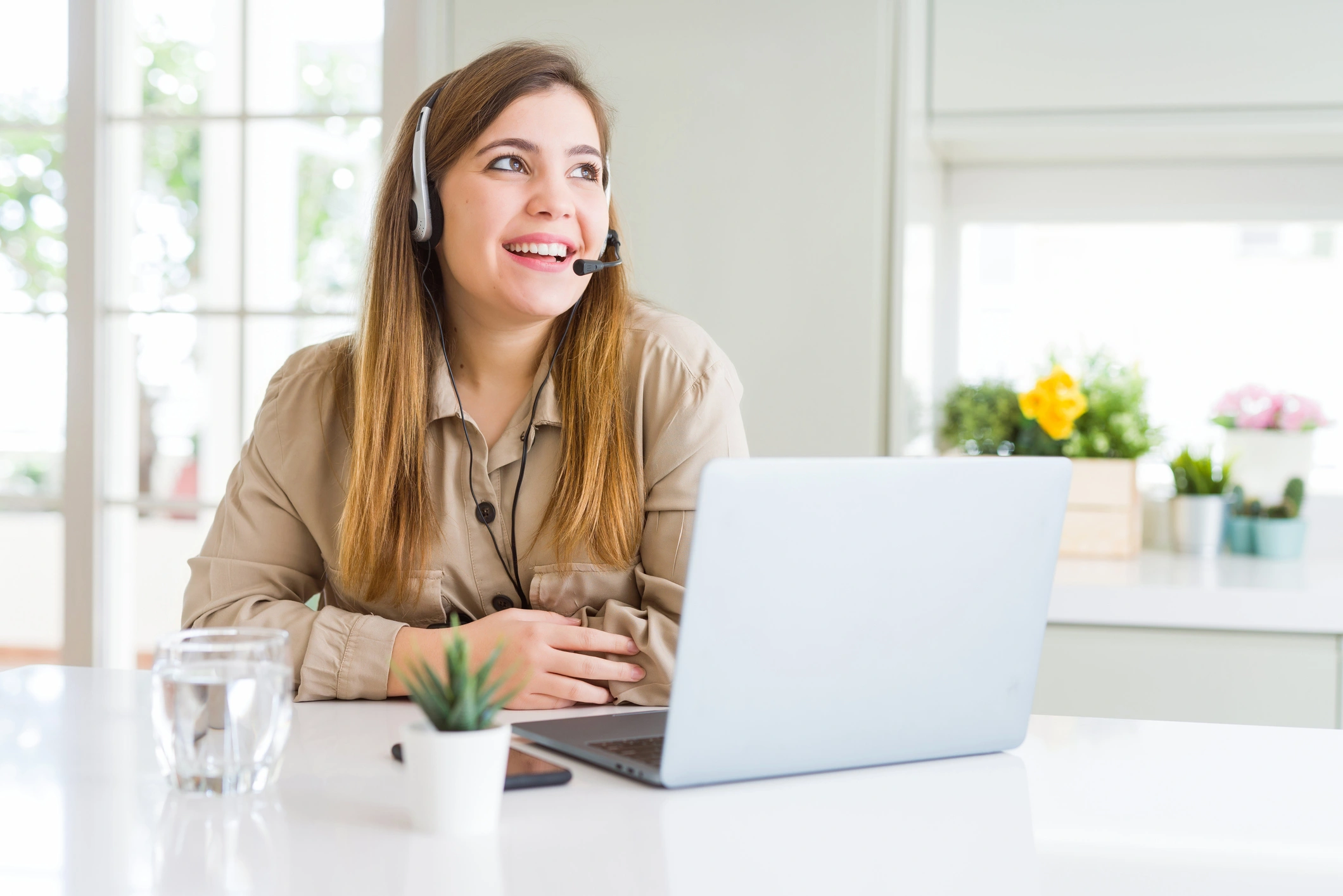 Smiling woman wearing a headset works on a laptop at a bright, modern desk.