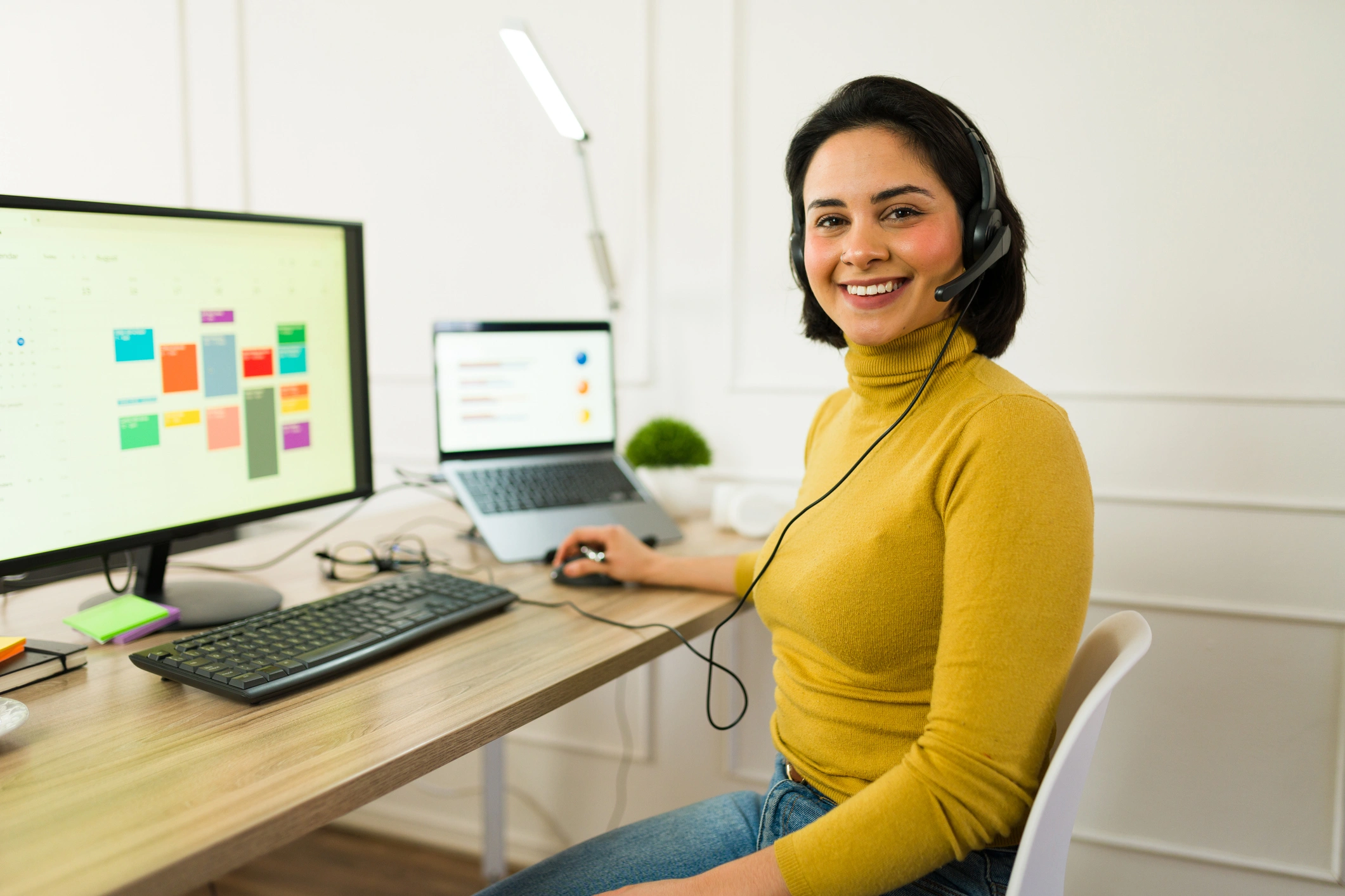 A woman in a yellow sweater smiles while working at a desk with a computer and laptop.