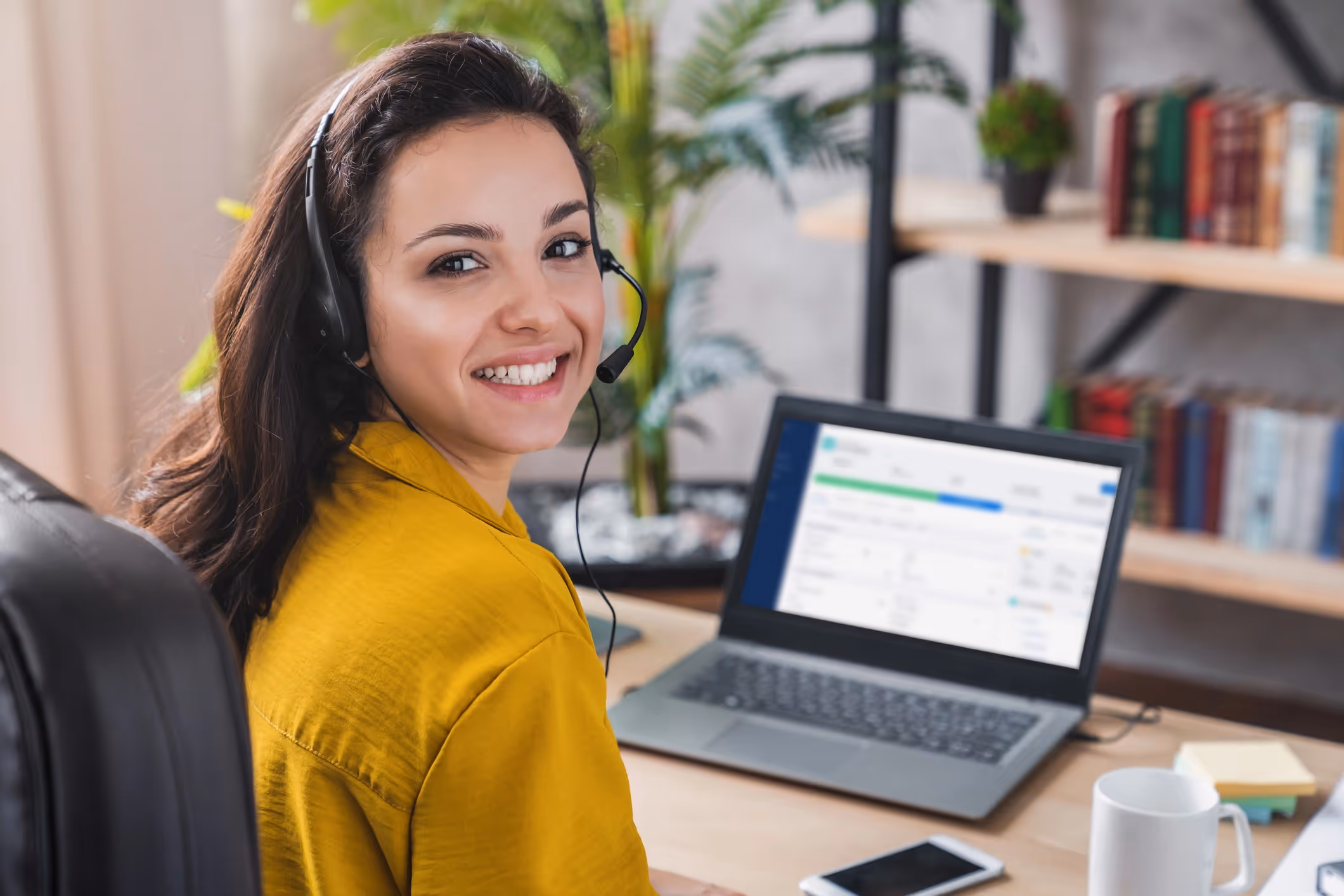 Smiling woman in a yellow shirt wearing a headset, sitting at a desk with an open laptop displaying work.