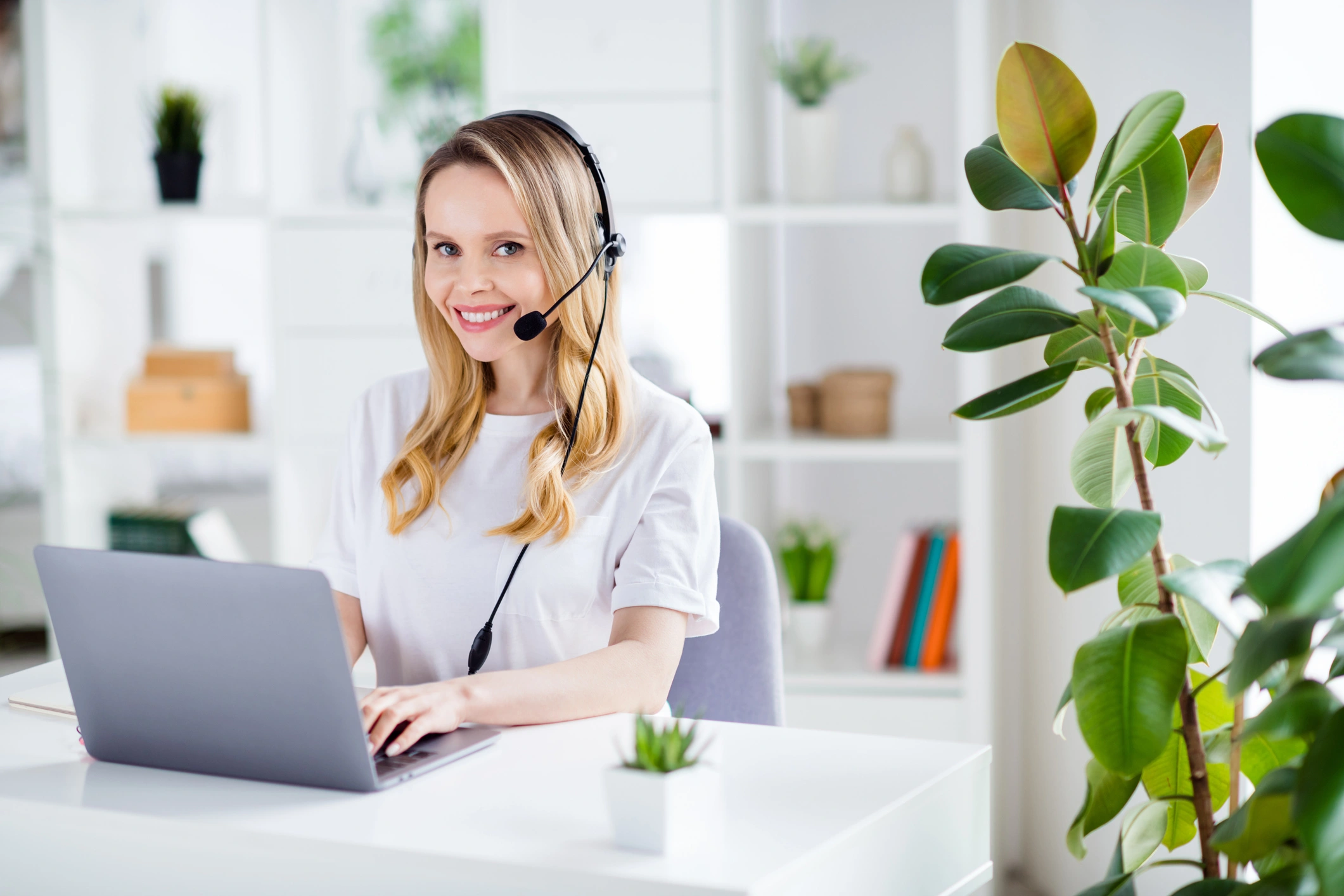 A woman with a headset sits smiling at a desk with a laptop, surrounded by plants. 