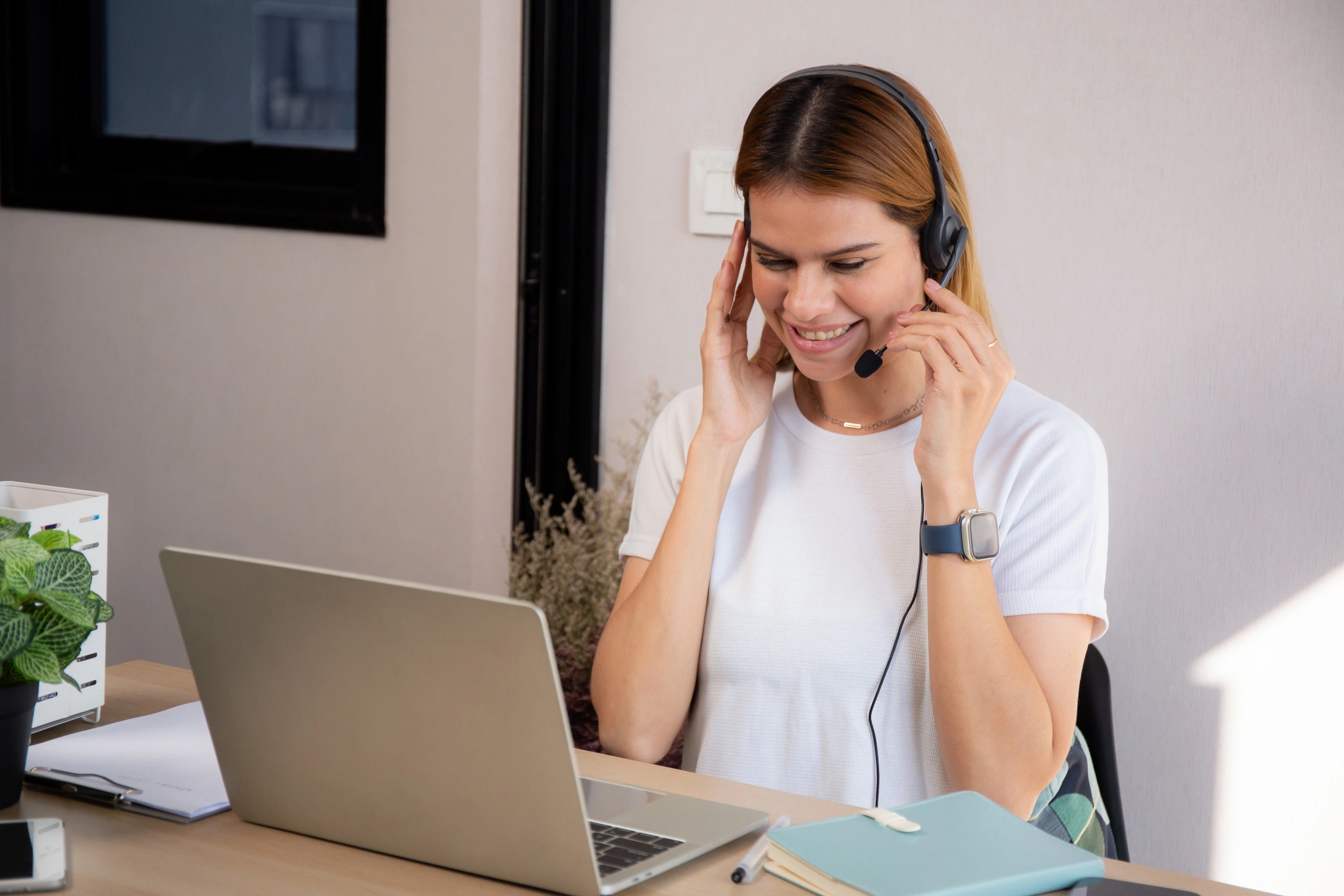 A woman in a white shirt smiles while using a headset and laptop.