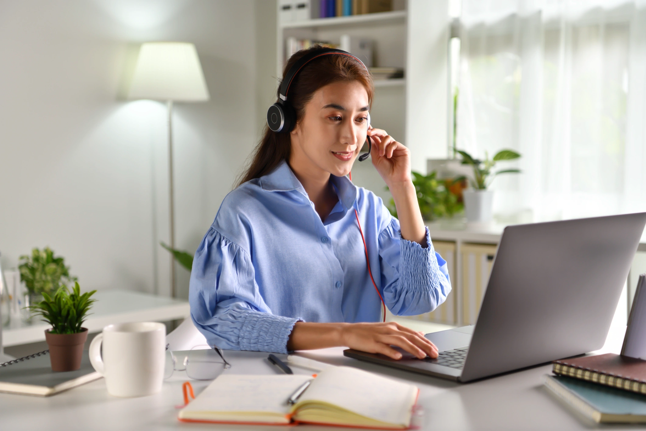 A woman in a blue blouse, wearing headphones, sits at a desk with a laptop, a notebook, and a mug. 