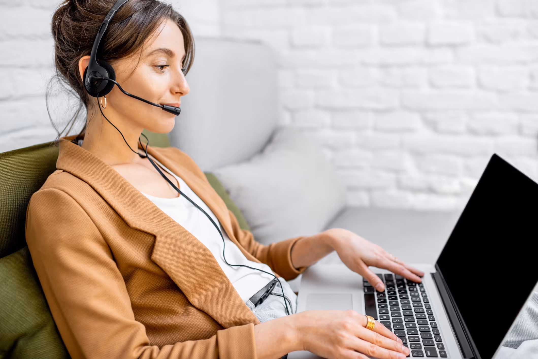A woman in a brown blazer uses a laptop while sitting on a couch, wearing a headset. 
