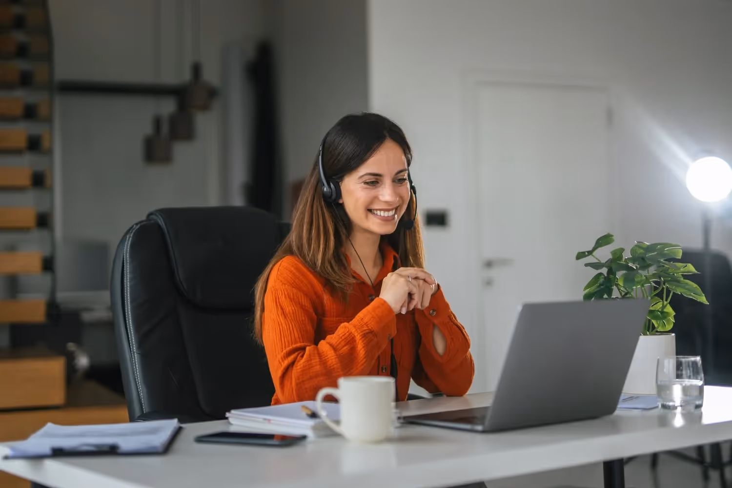  A smiling young woman with long dark hair is seated at a white desk in a home office, wearing a headset and an orange long-sleeved shirt. She is looking at her laptop screen, with her hands clasped, suggesting she is engaged in a video call or providing virtual customer support. A coffee mug, papers, and a potted plant are on the desk, with a staircase and a doorway visible in the background.