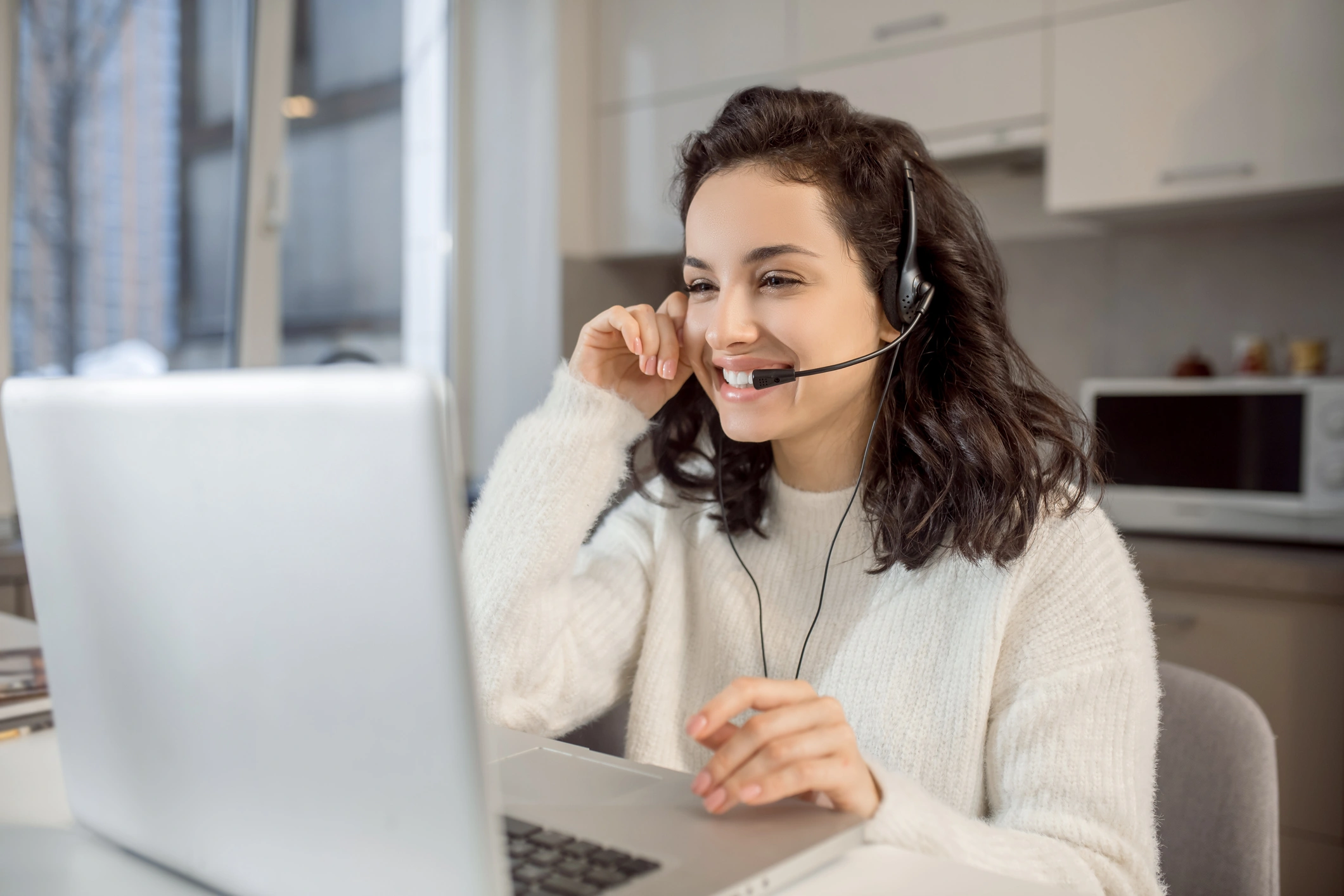 A woman in a white sweater, smiling and wearing a headset, sits in front of a laptop in a bright kitchen.