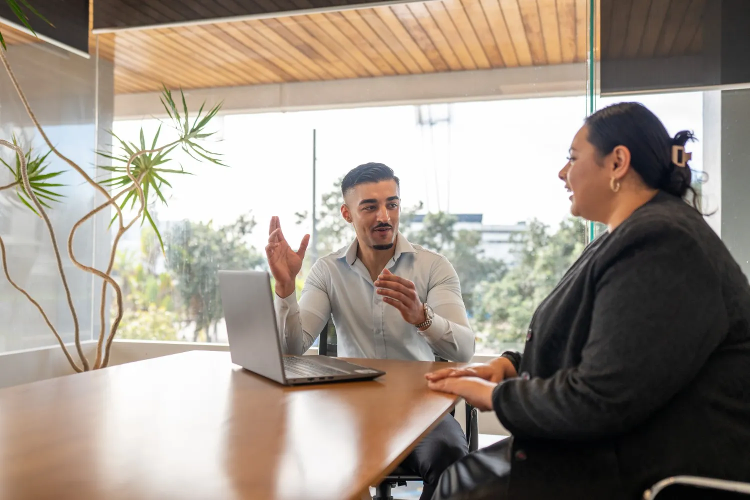 A man sits at a table with a laptop, gesturing expressively while talking to a woman in a professional office setting.