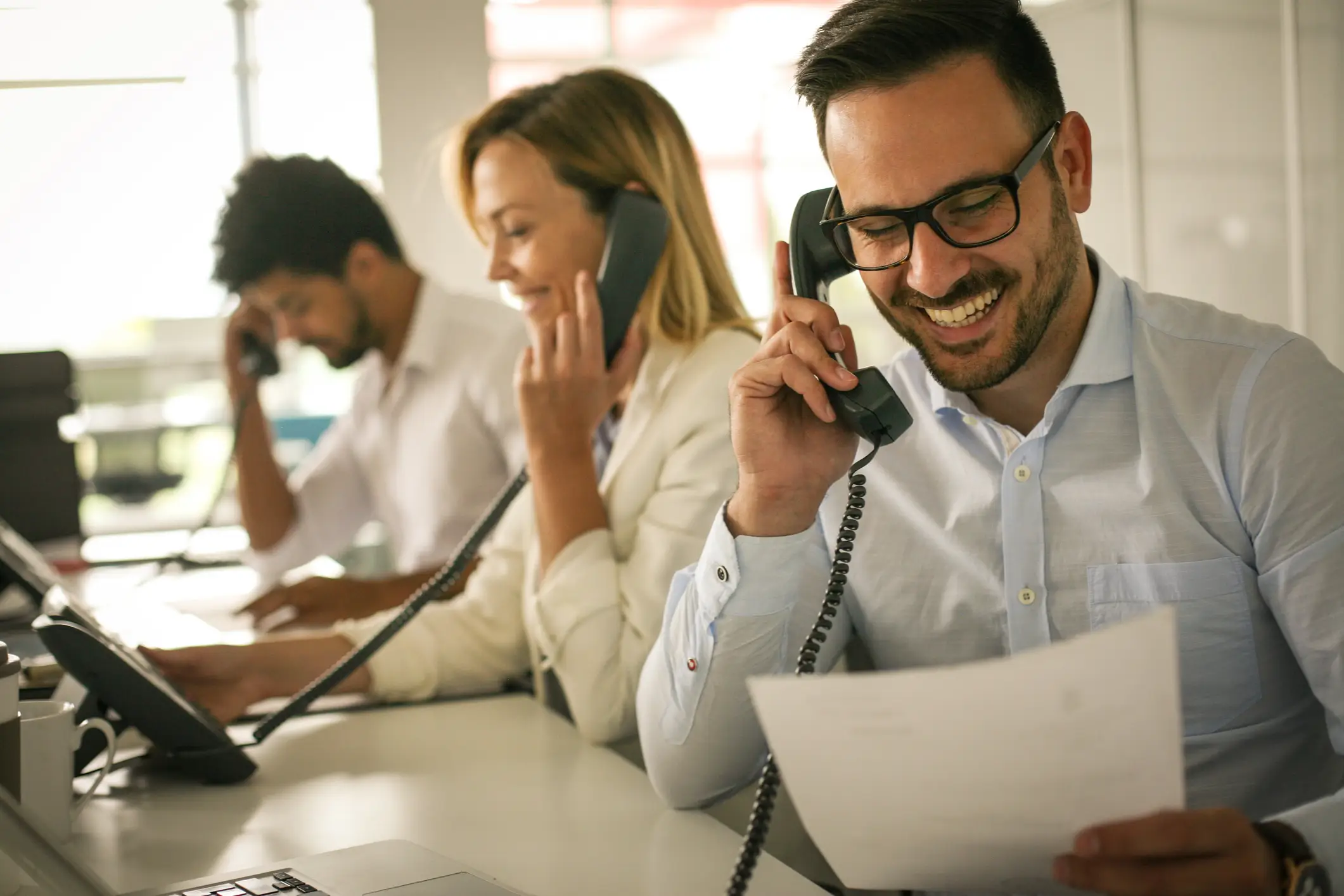 Cheerful businessman in glasses talking on the phone and holding paper, with coworkers nearby.