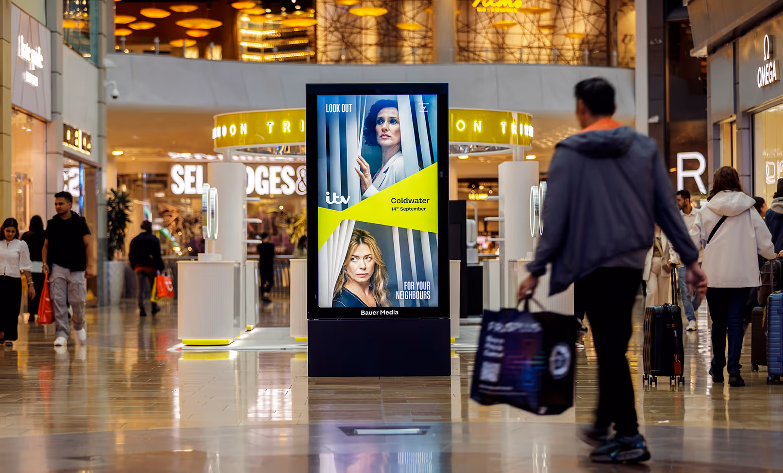 ITV advert plays on a mall digital advertising screen. People walk past with bags.