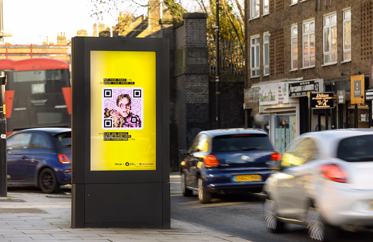 Cars driving on a street, there's a London bus. Ad advert is prominent for Elton John featuring a bespoke QR code to donate.
