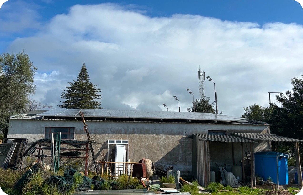 Rural building with solar panels on roof, various equipment outside, under a partly cloudy sky.