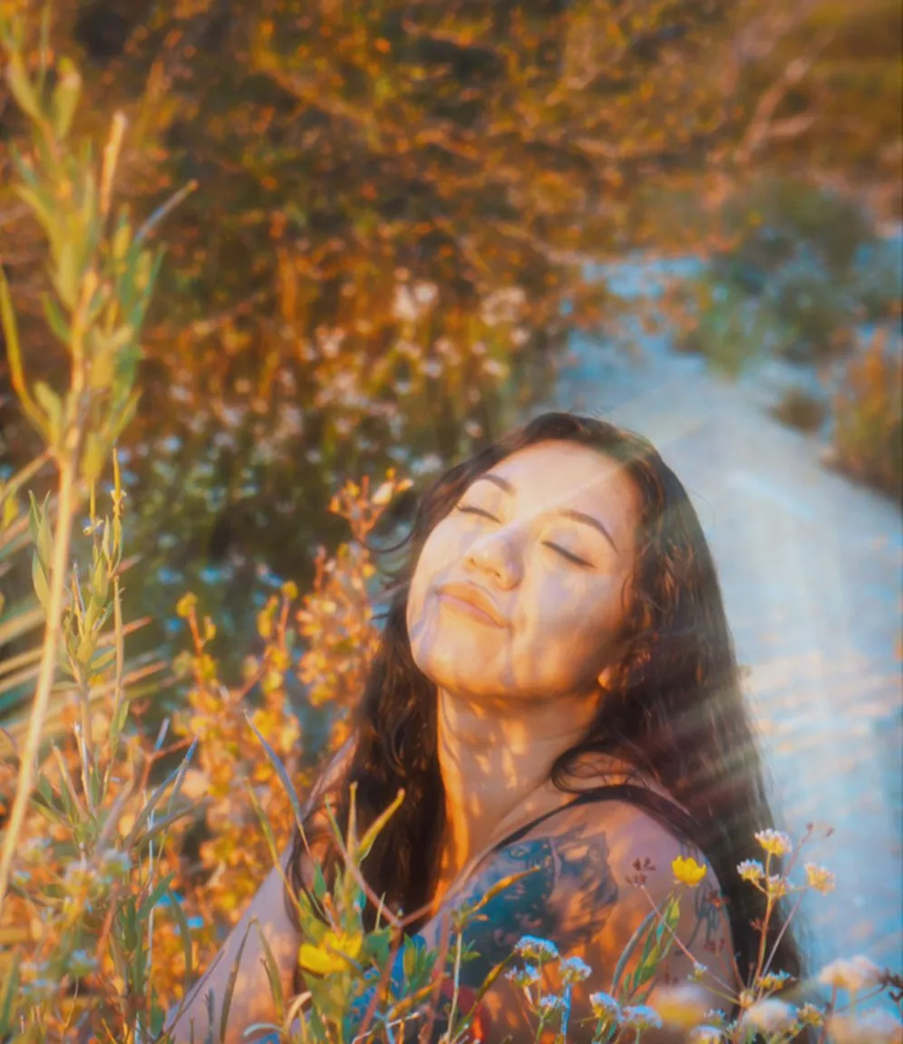 Woman with closed eyes and long dark hair enjoying sunlight surrounded by wildflowers and greenery.