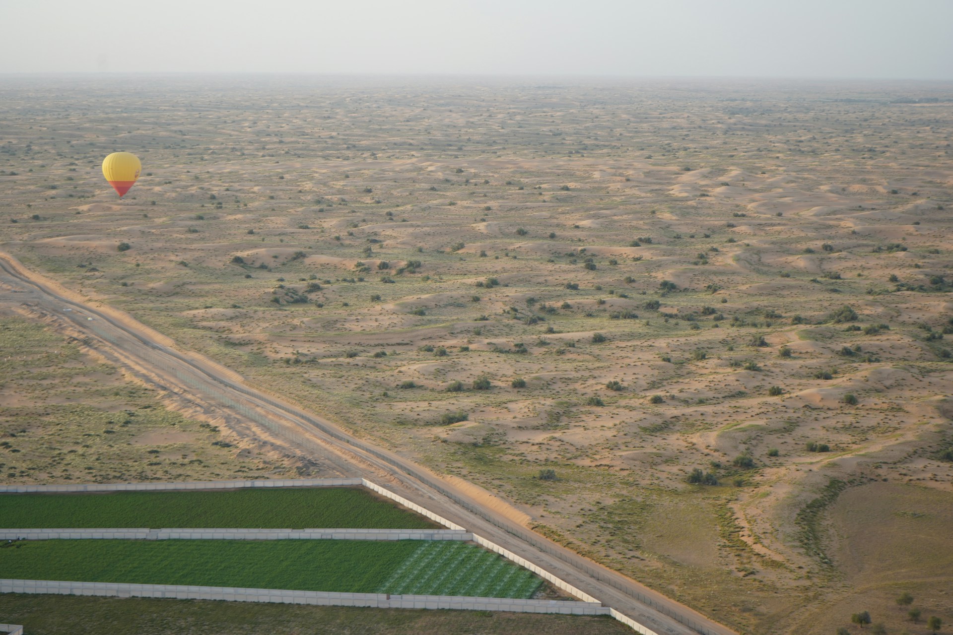 Aerial view of a desert landscape with patches of green agricultural fields and a yellow and red hot air balloon floating above.