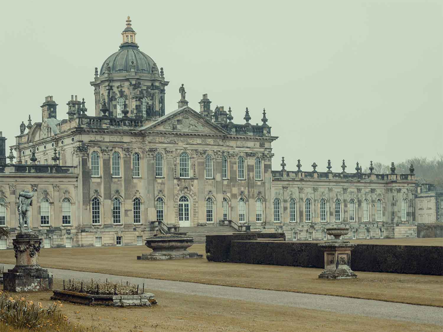 Large historic stone building with a central dome and ornate windows, set in a garden with statues and trimmed hedges.