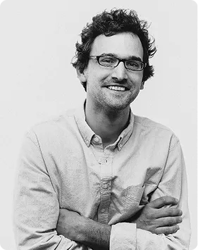 Mężczyzna w okularach i jasnej koszuli z rękawami podwiniętymi, uśmiechający się, z założonymi rękami.