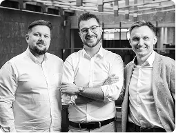 Uśmiechnięty mężczyzna w jasnej koszulce i ciemnej marynarce na jasnym tle.