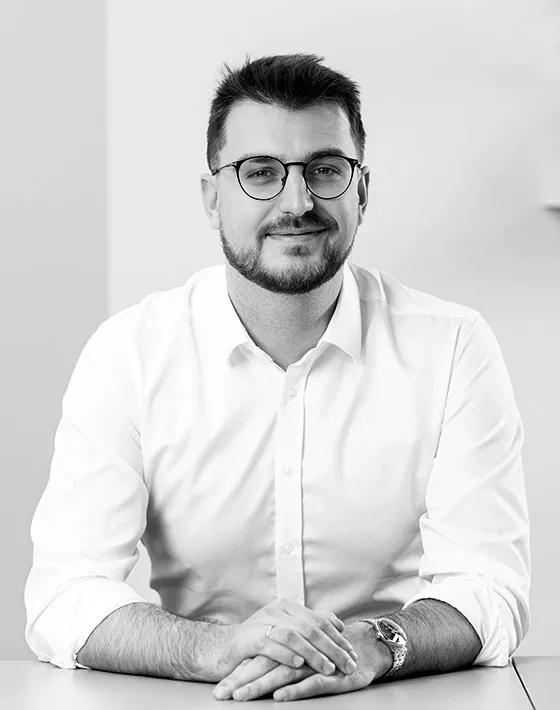 Mężczyzna w okularach i jasnej koszuli z rękawami podwiniętymi, uśmiechający się, z założonymi rękami.