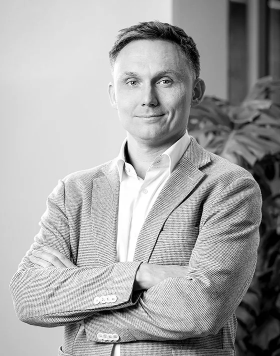 Mężczyzna w okularach i jasnej koszuli z rękawami podwiniętymi, uśmiechający się, z założonymi rękami.