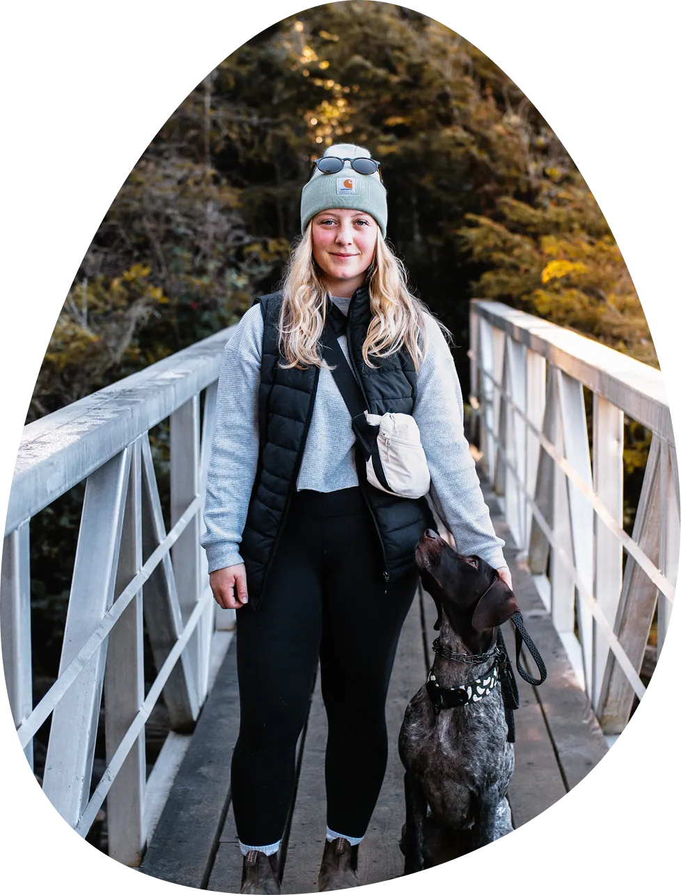 Woman wearing a beanie, sunglasses, and a black vest standing on a metal bridge holding a leash with a dog looking up at her.