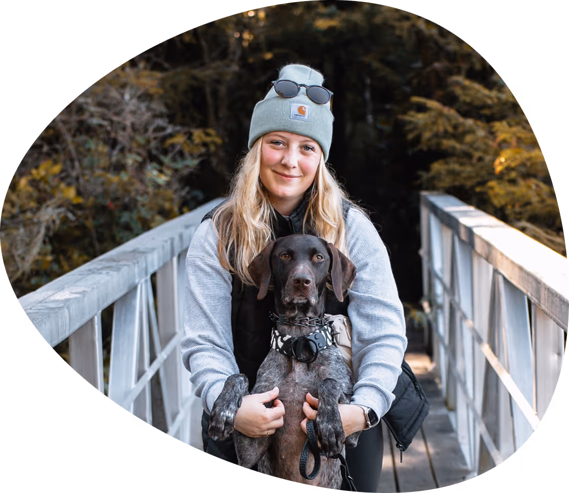 Woman wearing a gray beanie and sweater holding a black and gray dog on a wooden bridge with surrounding trees.