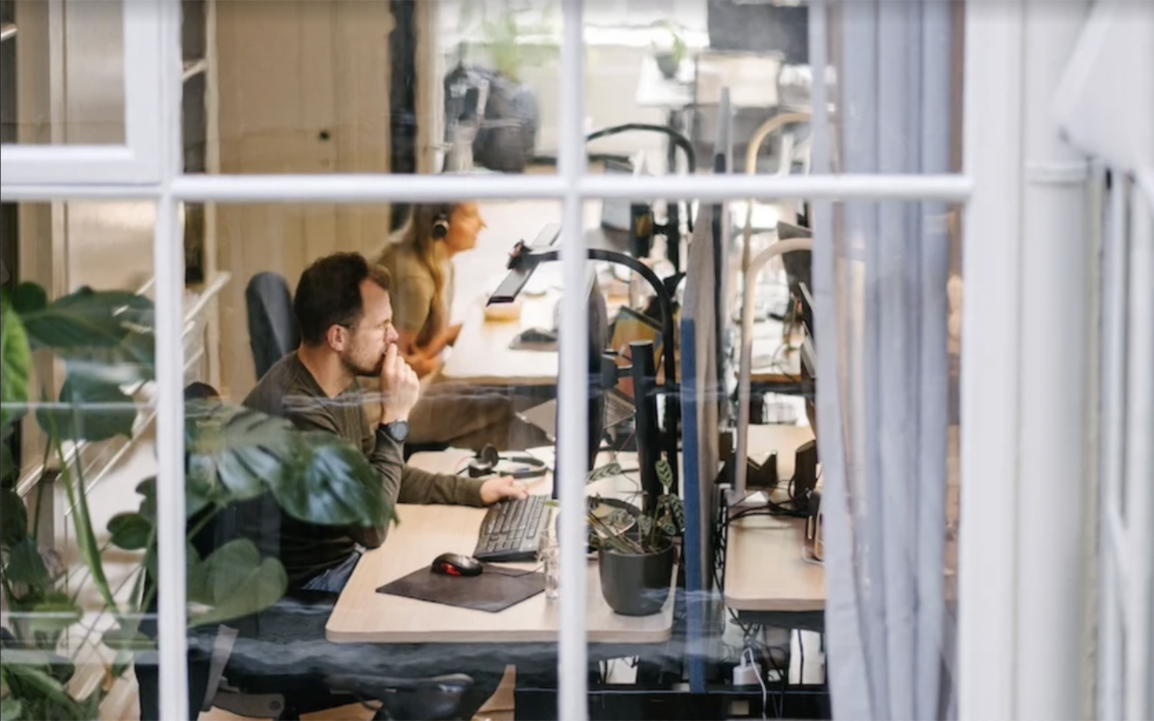 Mensen die geconcentreerd achter computers werken in een kantoorruimte, zichtbaar door een raam.