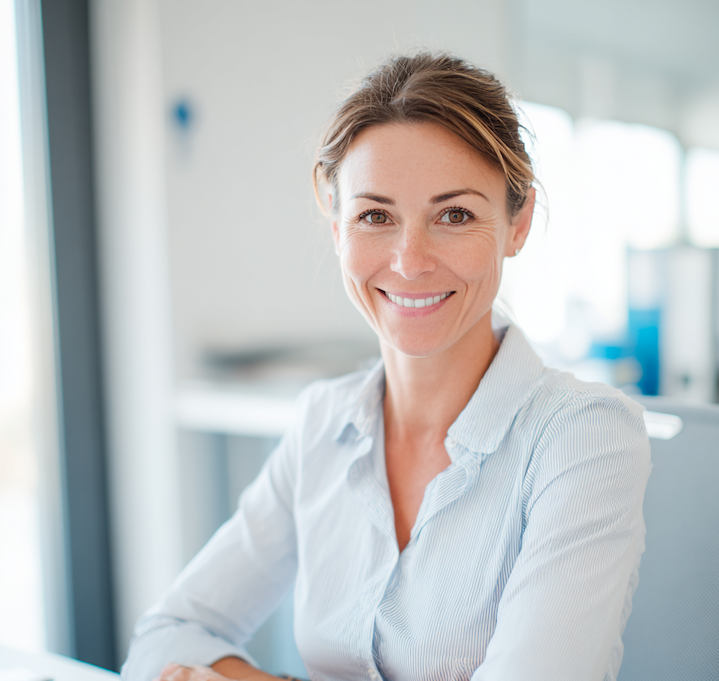 Vrouw met een blauwe blouse glimlacht zittend in een lichte kantooromgeving.