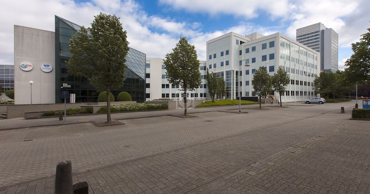Kantoorgebouw met glazen gevel en bomen langs een rustige straat onder een bewolkte lucht.