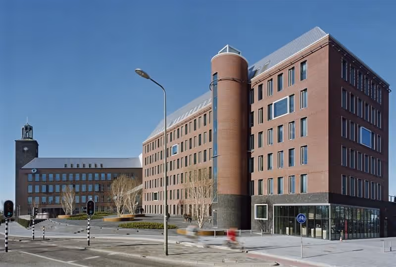 Modern kantoorpand met schuin dak, ronde toren aan de voorkant en veel ramen, gelegen aan een kruispunt met verkeerslichten.