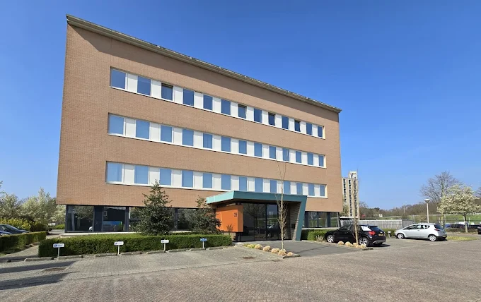 Modern kantoorgebouw met vier verdiepingen en parkeerplaatsen eromheen onder een blauwe lucht.