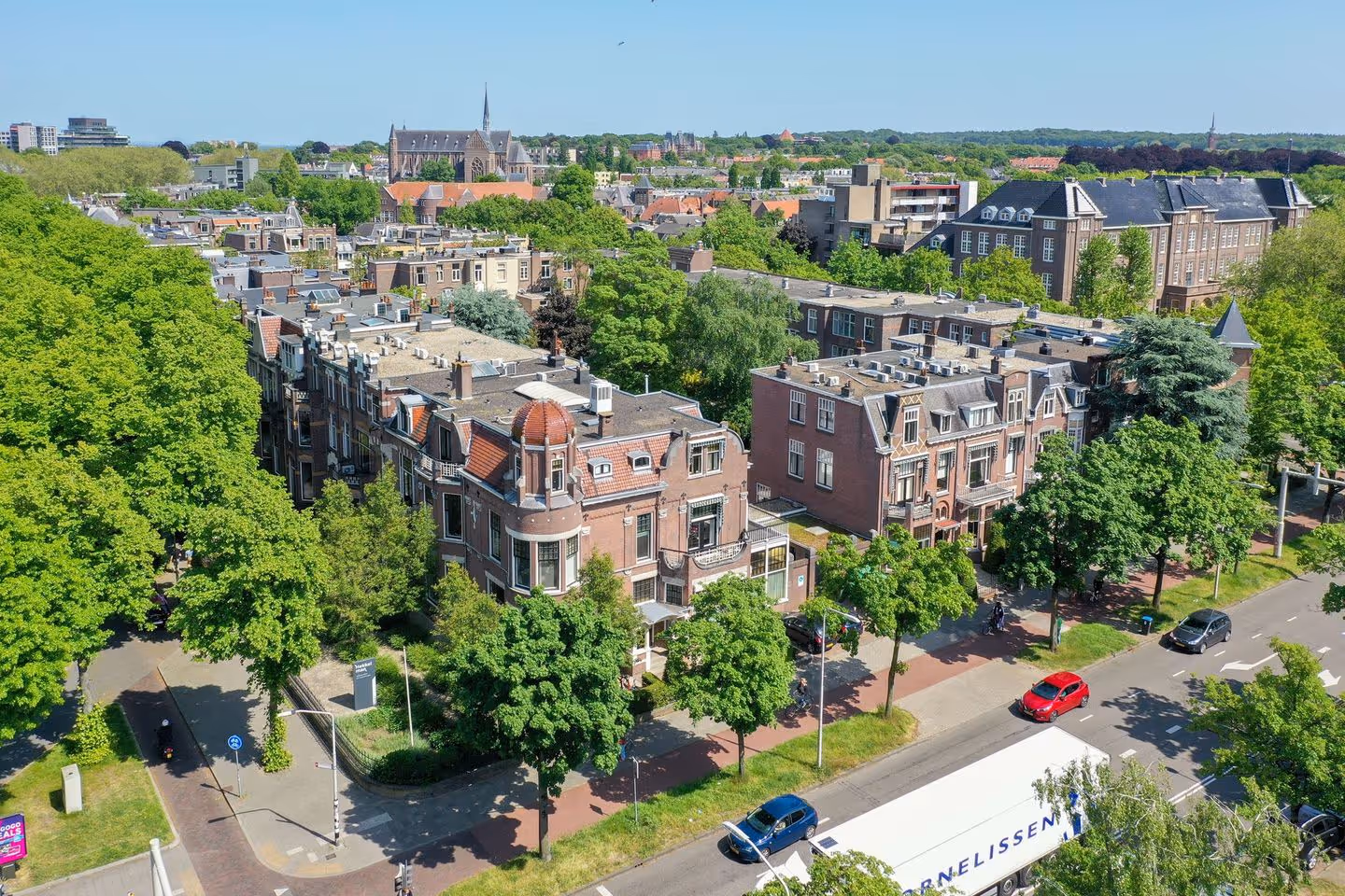 Luchtfoto van een groene, stedelijke buurt met historische gebouwen, bomen en een straat met auto's.