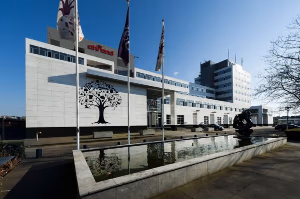 Modern kantoorgebouw met witte gevel, vlaggenmasten en een langwerpige fontein op een zonnige dag.