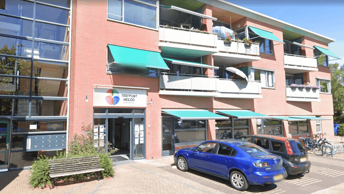 Modern gebouw met rode bakstenen en balkon met groene luifels, parkeerplaatsen met geparkeerde auto's en fietsenrekken.