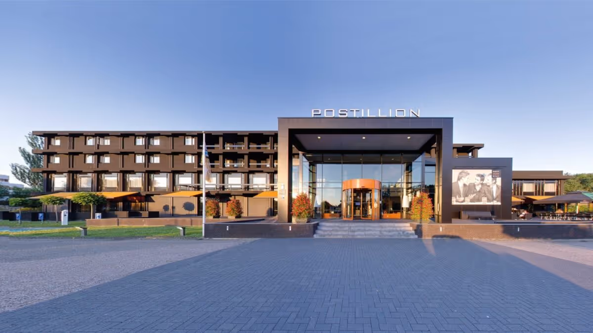 Vooraanzicht van het moderne Postillion hotelgebouw met glazen ingang en terrassen onder oranje parasols.