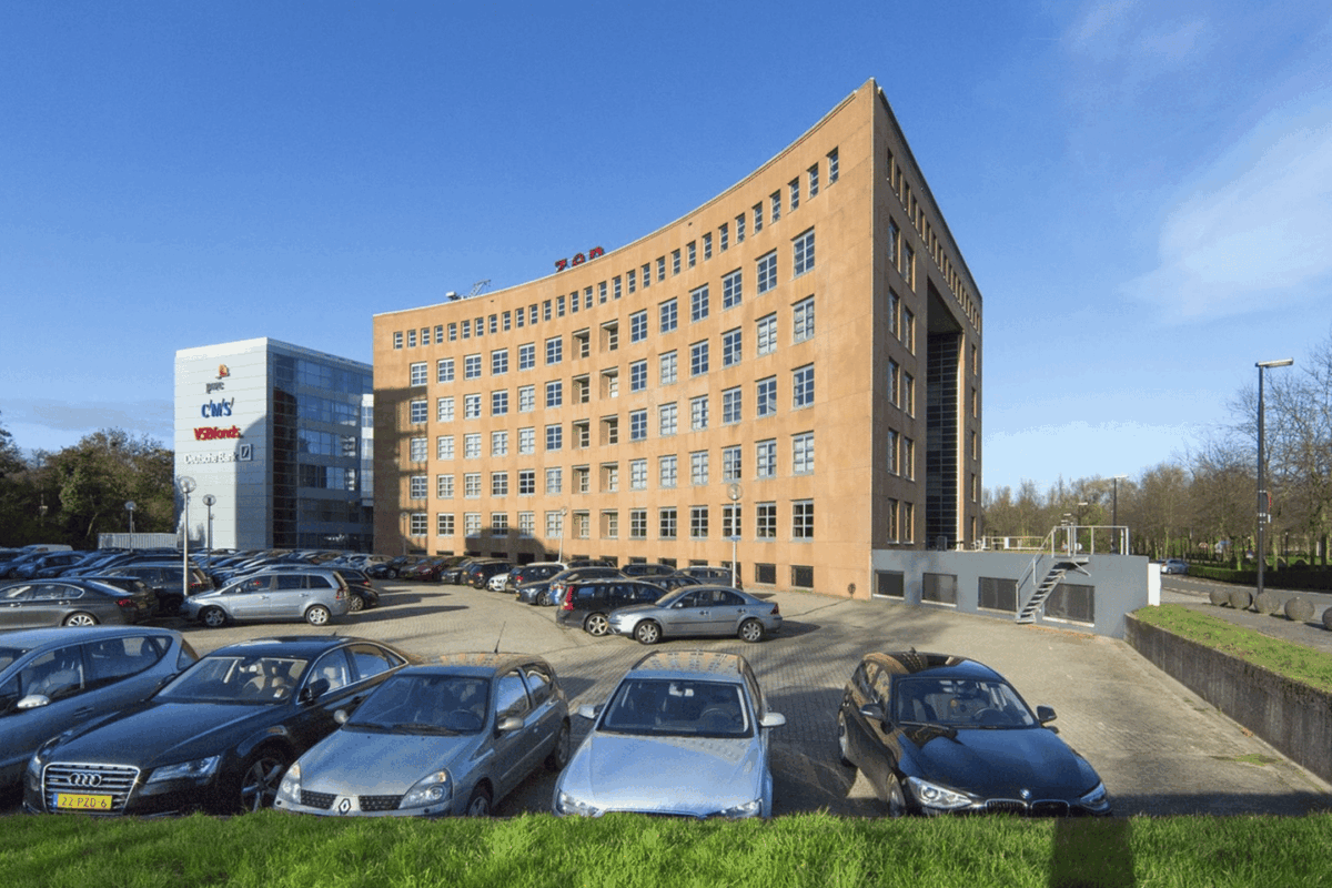 Parkeerterrein met geparkeerde auto's voor een groot kantoorgebouw onder een blauwe lucht.