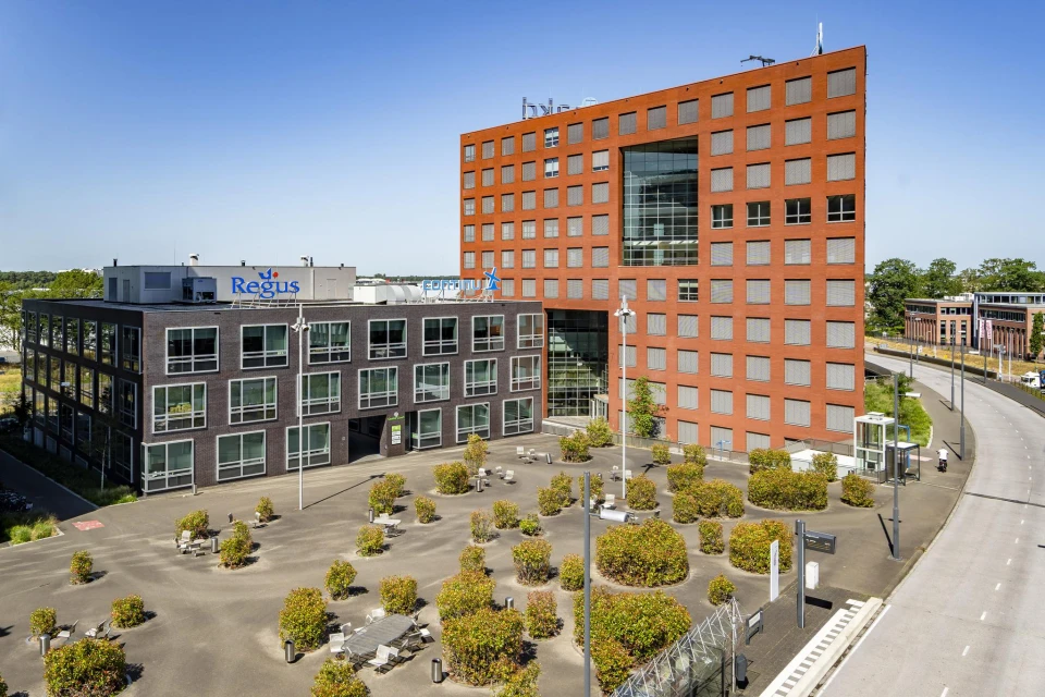 Modern kantoorgebouw met twee verbonden delen, omgeven door een plein met verspreide groene struiken en tafels met stoelen.