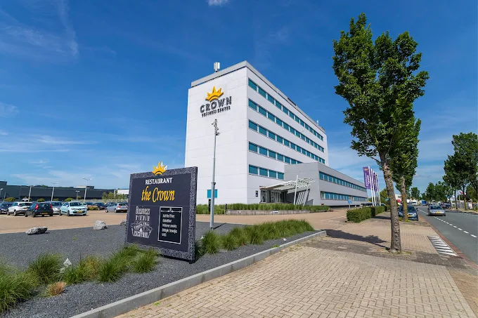 Modern wit kantoorgebouw met het logo van Crown Restaurant en een bord met het menu aan de voorkant langs een straat met bomen en een parkeerplaats onder een blauwe lucht.