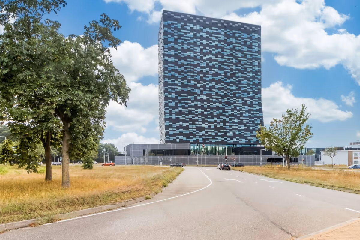 Modern hoog gebouw met een patroon van licht- en donkerblauwe ramen, gezien vanaf een lege weg met bomen aan de zijkant.