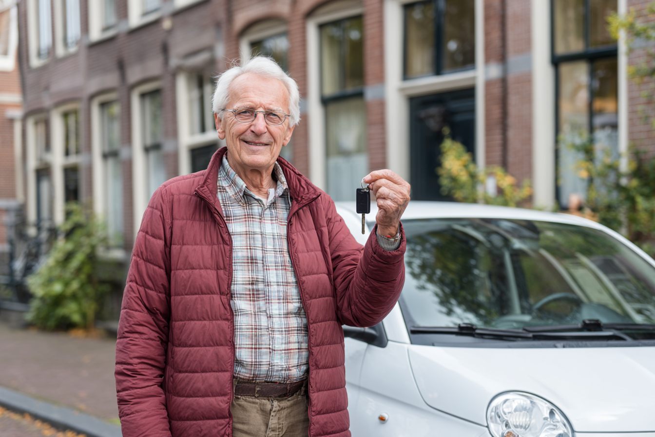 Oudere man met grijs haar en bril staat buiten naast een witte auto en houdt een autosleutel omhoog.