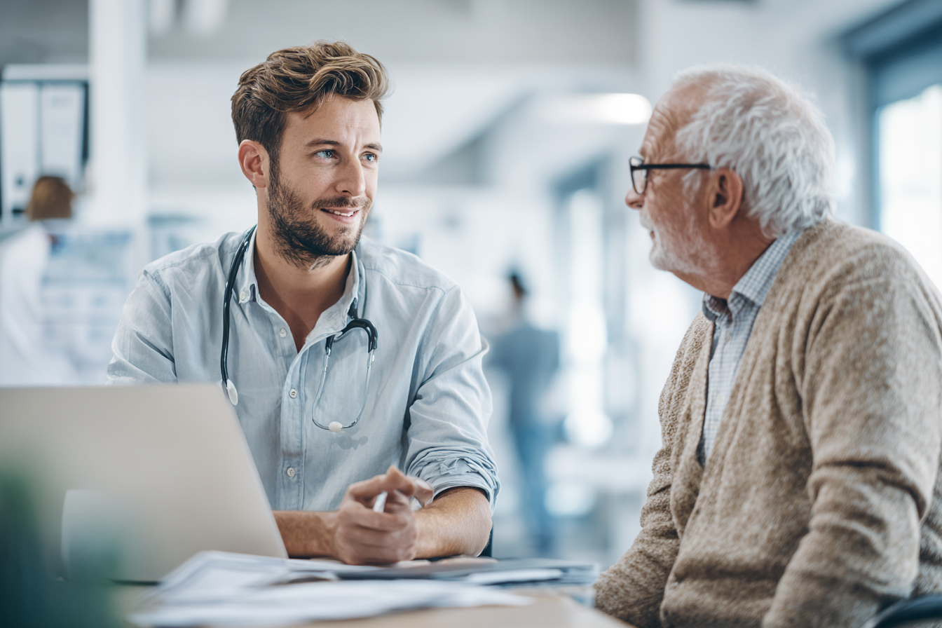 Jonge mannelijke arts met stethoscoop praat met een oudere man in een lichte medische omgeving.