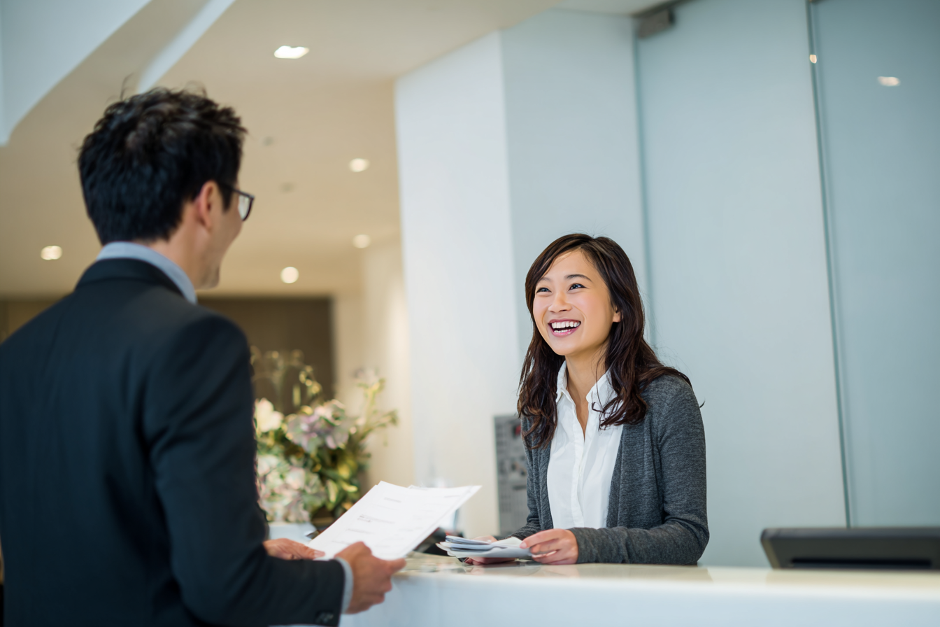 Lachende vrouw achter balie helpt een man in een pak met documenten in een kantooromgeving.