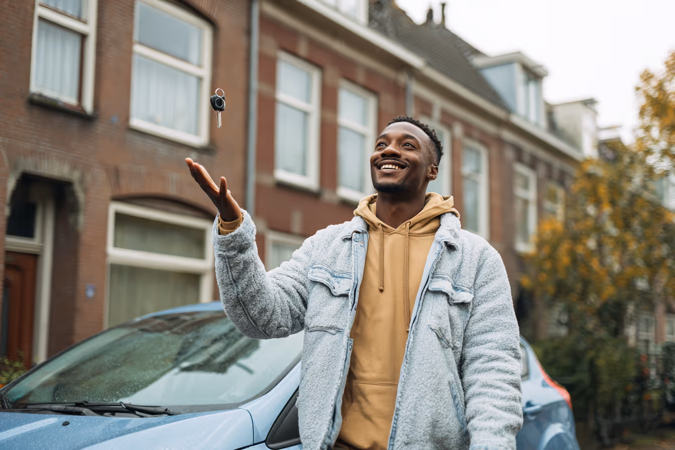 Man gooit autossleutel in de lucht staand naast een blauwe auto in een woonstraat.