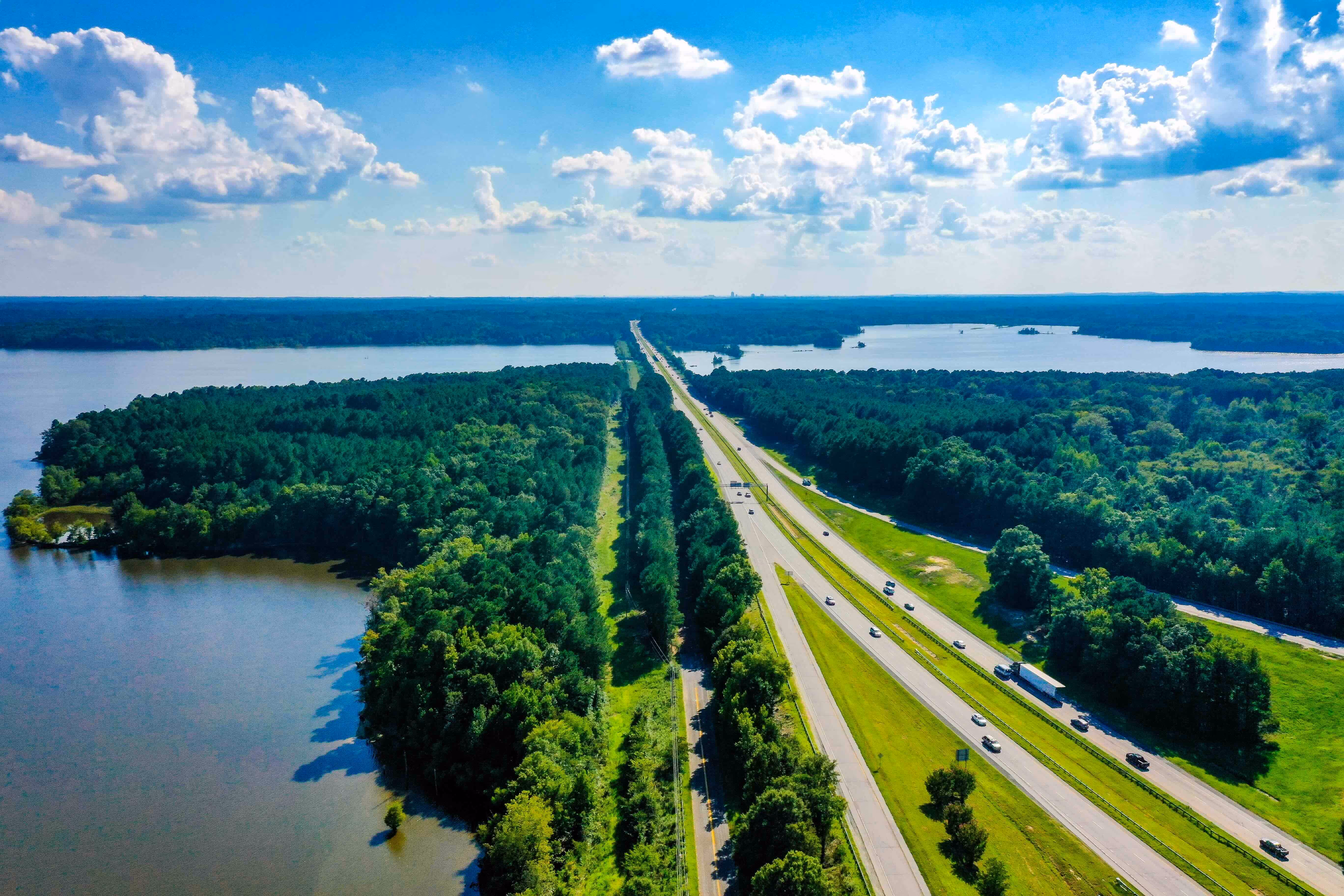 Luchtfoto van snelweg met verkeer, omgeven door groen beboste gebieden en een groot meer onder een blauwe lucht met wolken.