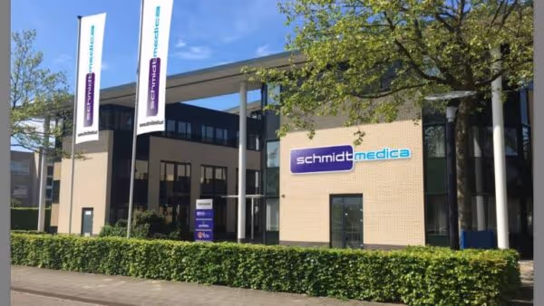 Kantoorgebouw met het logo van Schmidt Medica op de gevel, omringd door groene hagen en vlaggen met bedrijfsnaam onder een blauwe lucht.