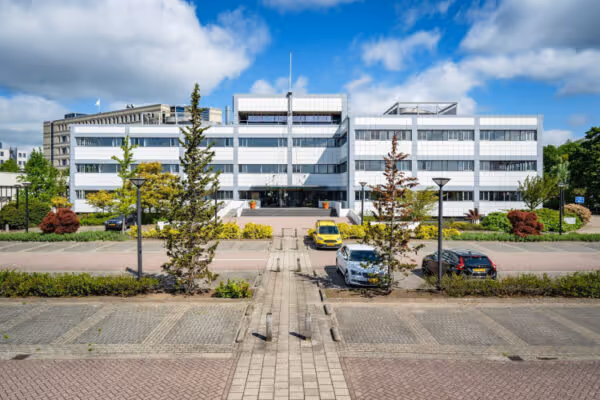 Modern wit kantoorgebouw met ramen, straat ervoor en een paar geparkeerde auto's onder een partly bewolkte blauwe lucht.