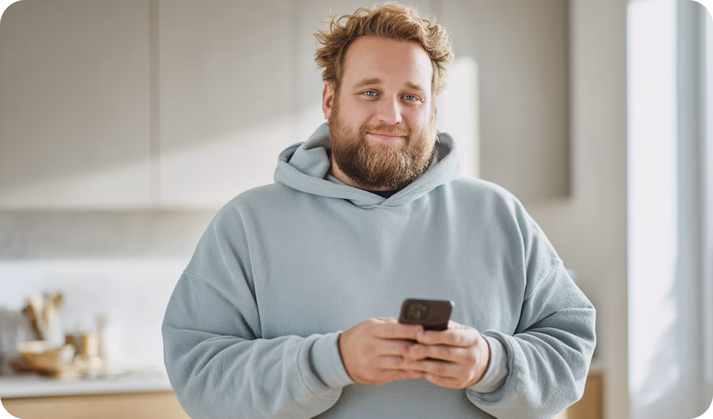 A man with a beard and light brown hair, wearing a light gray hoodie, smiles while holding a smartphone in both hands standing in a bright, modern kitchen.