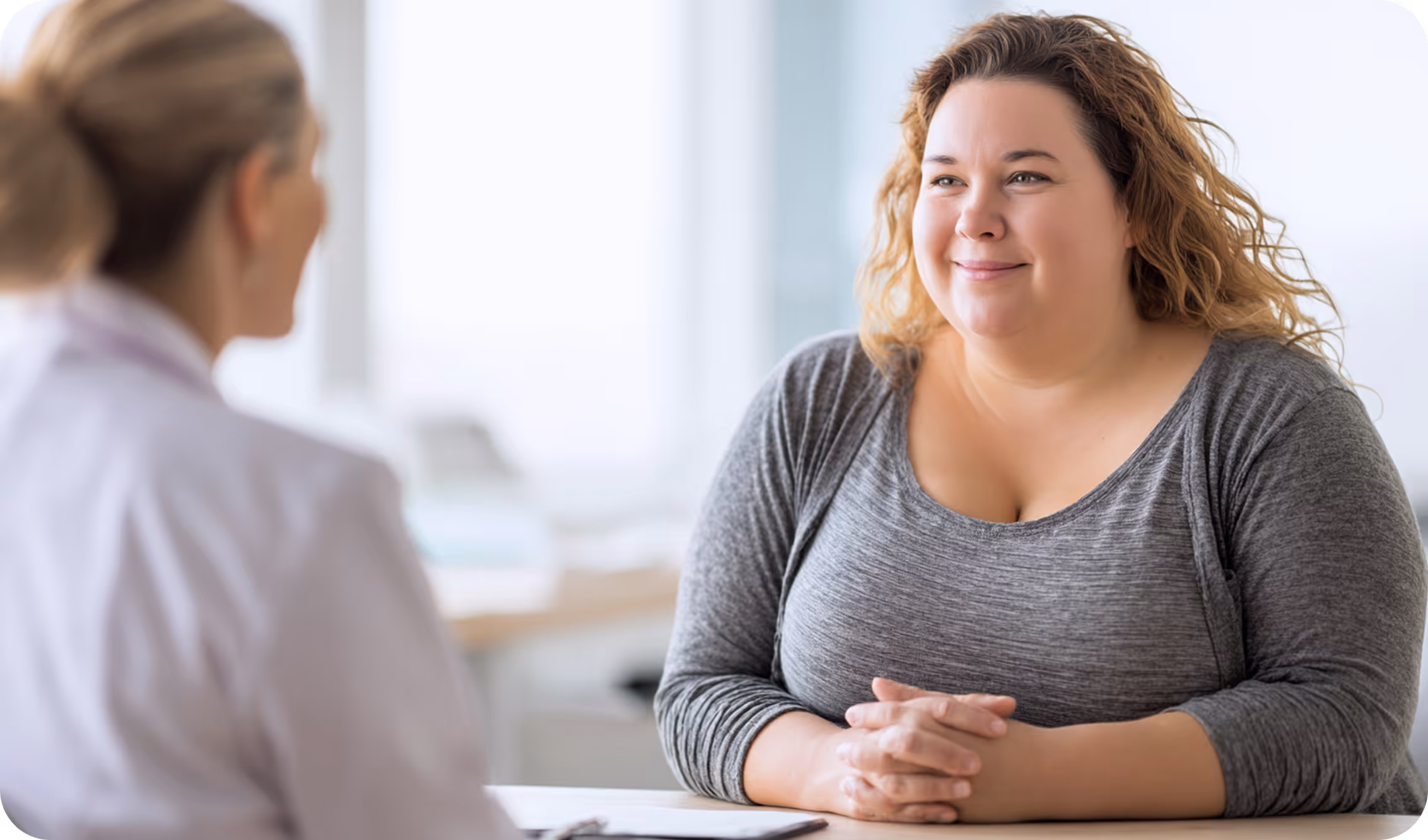 A woman with long, curly hair smiles while sitting at a desk across from a healthcare professional in a bright, clinical setting.