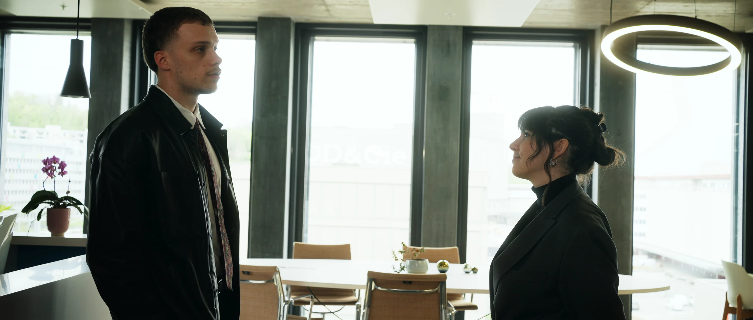 Un homme en blouson de cuir noir et une femme se font face en contre-jour devant de grandes baies vitrées, sous une suspension circulaire lumineuse, dans un espace de bureaux.