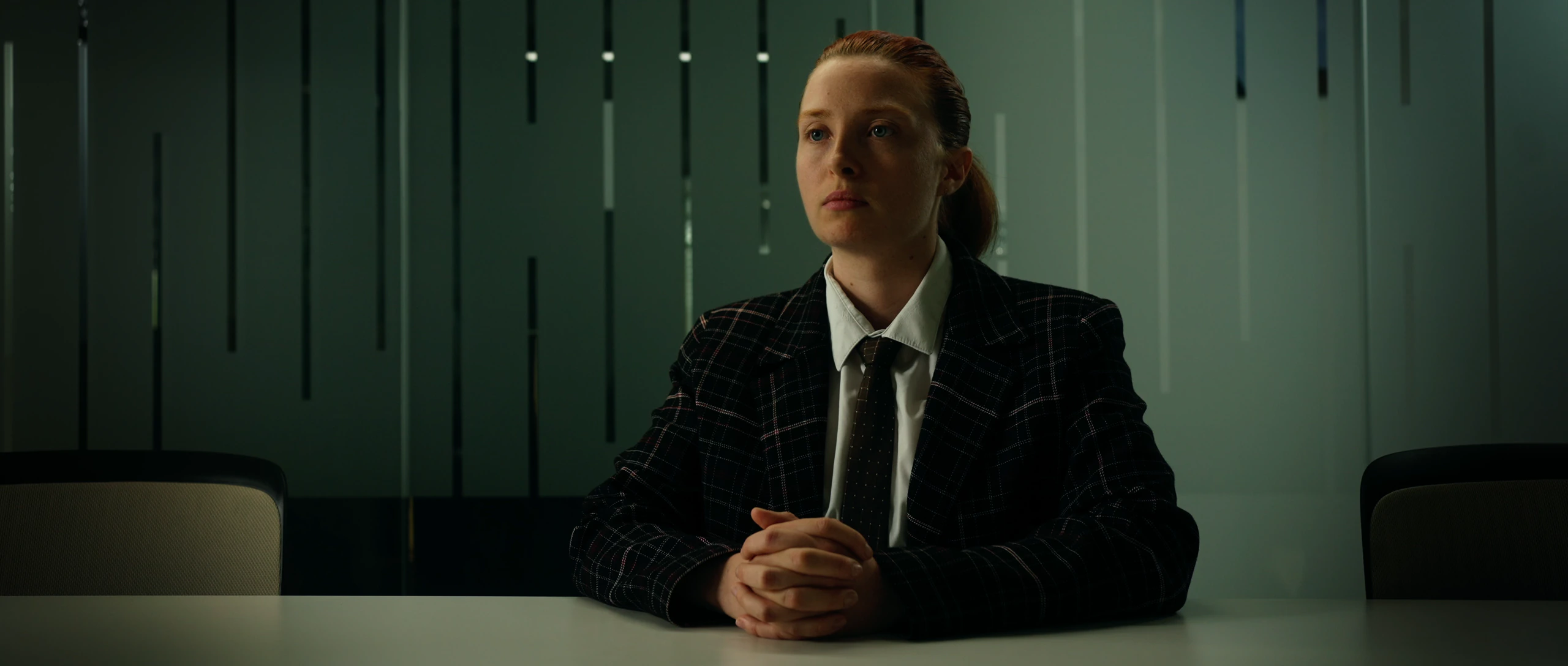 Une jeune femme rousse en veste à carreaux, assise les mains jointes à une table de réunion, devant une cloison vitrée verte.
